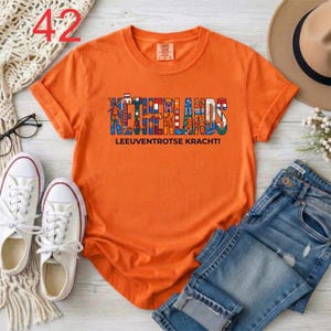 Puede incluir: Una camiseta naranja de manga corta con un dise&ntilde;o gr&aacute;fico que muestra la palabra NETHERLANDS en letras grandes y coloridas rellenas de ilustraciones holandesas, y el texto LEEUVENTROTSE KRACHT! impreso en negro bajo el gr&aacute;fico.