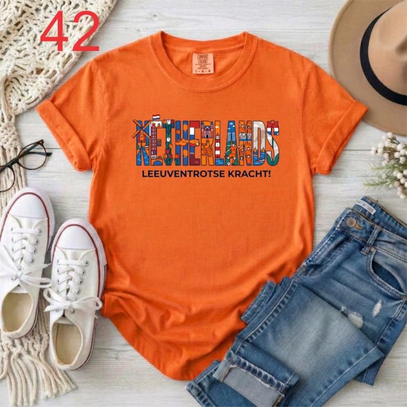 Puede incluir: Una camiseta naranja de manga corta con un dise&ntilde;o gr&aacute;fico que muestra la palabra NETHERLANDS en letras grandes y coloridas rellenas de ilustraciones holandesas, y el texto LEEUVENTROTSE KRACHT! impreso en negro bajo el gr&aacute;fico.