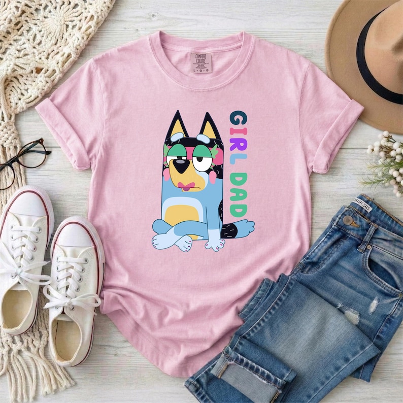 May include: A light pink t-shirt featuring a graphic of a blue and yellow cartoon dog sitting with legs crossed. The text "GIRL DAD" is printed vertically in colorful letters next to the dog. The shirt has a crew neckline and short sleeves.