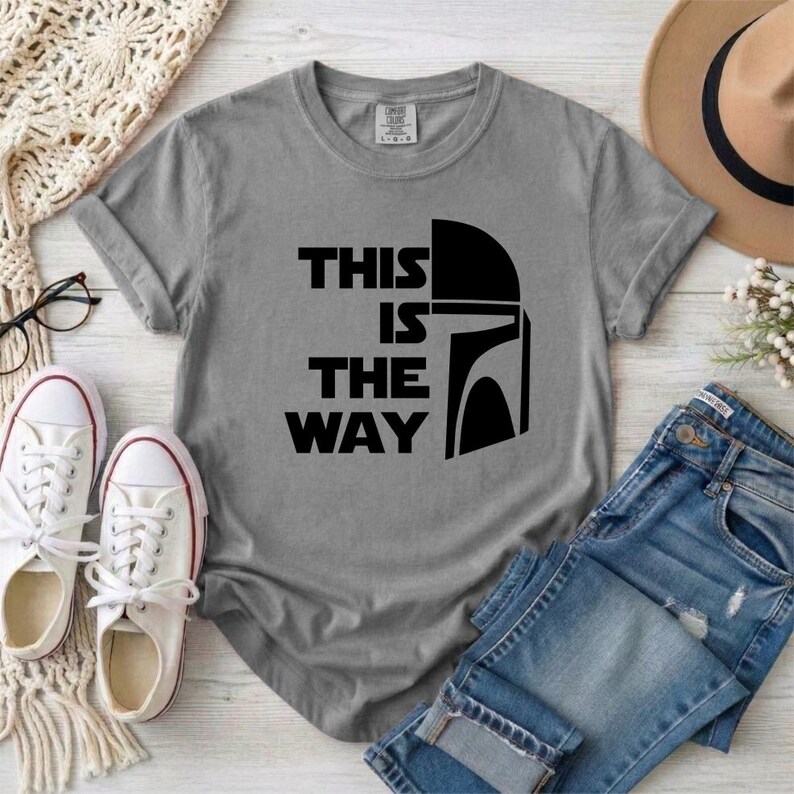May include: Gray t-shirt with the text "THIS IS THE WAY" and a black helmet graphic. The shirt is paired with blue jeans, white sneakers, and a brown hat. The shirt is a comfortable, casual style.