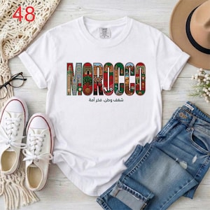Puede incluir: Una camiseta blanca de manga corta con la palabra MOROCCO en letras grandes y coloridas, cada una rellena con patrones intrincados e ilustraciones de monumentos marroqu&iacute;es. Debajo del texto aparece la frase en &aacute;rabe شغف وطن، فخر أمة.