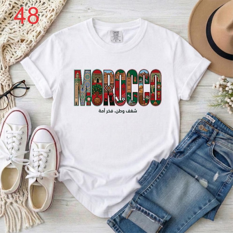 Puede incluir: Una camiseta blanca de manga corta con la palabra MOROCCO en letras grandes y coloridas, cada una rellena con patrones intrincados e ilustraciones de monumentos marroqu&iacute;es. Debajo del texto aparece la frase en &aacute;rabe شغف وطن، فخر أمة.