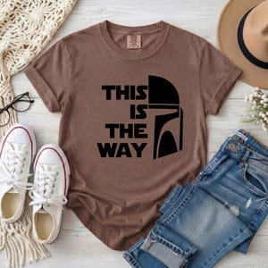 May include: A brown t-shirt with the text "THIS IS THE WAY" and a black helmet graphic. The shirt is paired with blue jeans, white sneakers, and a tan hat. A cream-colored scarf and eyeglasses are also visible.