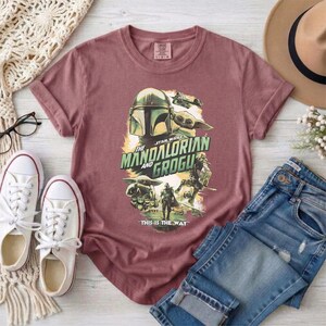 May include: A maroon t-shirt featuring a graphic from "The Mandalorian" with the text "The Mandalorian and Grogu" and the phrase "This is the Way." The design includes images of the Mandalorian, Grogu, and other characters from the show.