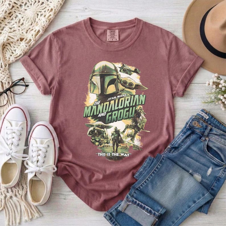 May include: A maroon t-shirt featuring a graphic from "The Mandalorian" with the text "The Mandalorian and Grogu" and the phrase "This is the Way." The design includes images of the Mandalorian, Grogu, and other characters from the show.