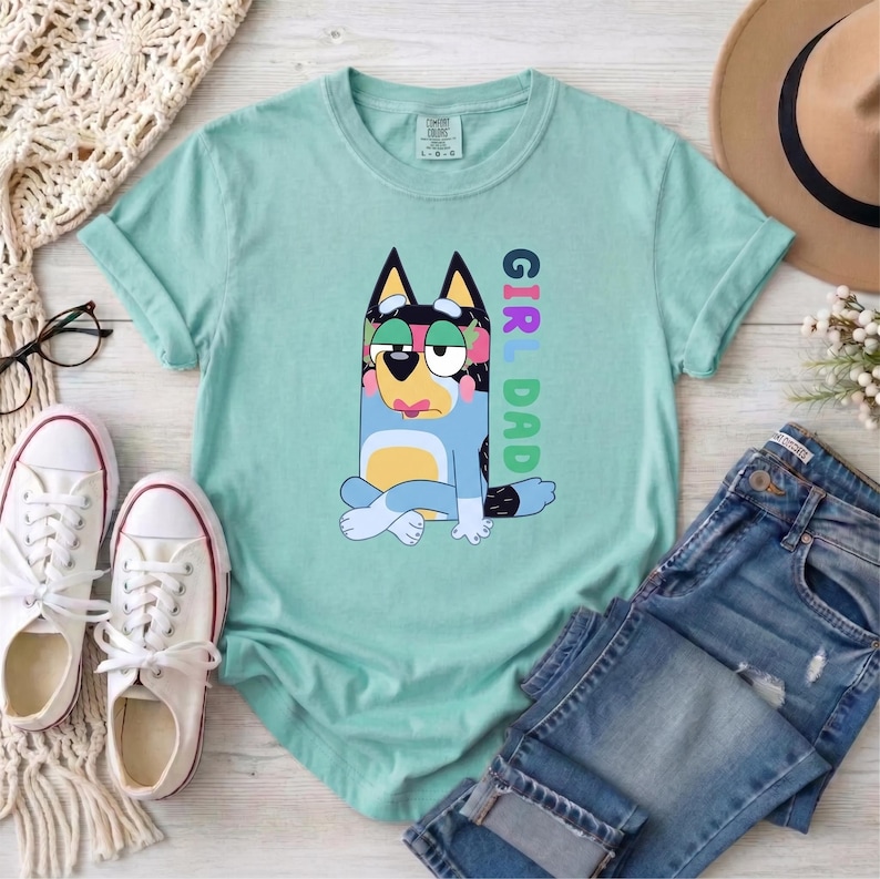 May include: A light mint green short-sleeve t-shirt featuring a graphic of a blue and yellow cartoon dog sitting with its legs crossed. The text "GIRL DAD" is printed vertically in colorful block letters next to the dog illustration.