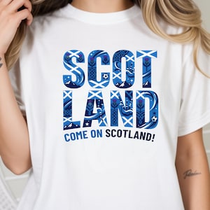 May include: White t-shirt with a blue and white graphic that says "SCOTLAND" in a decorative font. The graphic includes thistle and soccer ball designs. Below the word "SCOTLAND" is the phrase "COME ON SCOTLAND!"