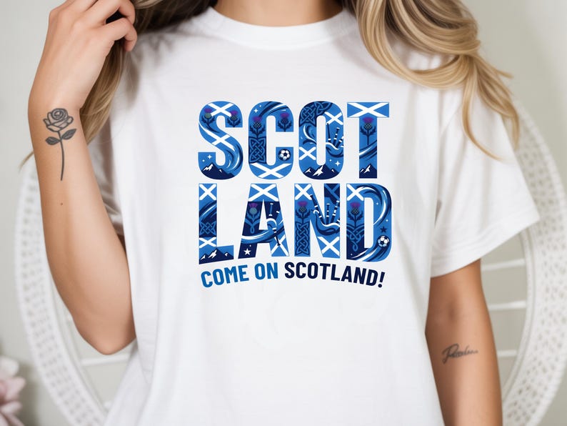 May include: White t-shirt with a blue and white graphic that says "SCOTLAND" in a decorative font. The graphic includes thistle and soccer ball designs. Below the word "SCOTLAND" is the phrase "COME ON SCOTLAND!"