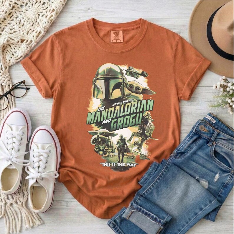 May include: An orange t-shirt featuring a graphic from "The Mandalorian" with the text "The Mandalorian and Grogu" and "This is the Way." The shirt is paired with blue jeans, white sneakers, and a beige hat.