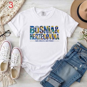 May include: White t-shirt with the words "BOSNIA & HERZEGOVINA" in colorful letters. Below the text is the phrase "NA&Scaron;I ZMAJEVI, NA&Scaron; PONOS!" in blue. The shirt is paired with blue jeans and white sneakers.