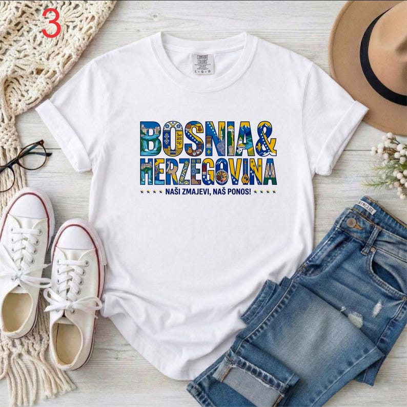 May include: White t-shirt with the words "BOSNIA & HERZEGOVINA" in colorful letters. Below the text is the phrase "NA&Scaron;I ZMAJEVI, NA&Scaron; PONOS!" in blue. The shirt is paired with blue jeans and white sneakers.