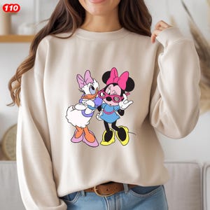 Pode incluir: Camisola bege com um gr&aacute;fico colorido da Minnie Mouse e da Margarida. Minnie Mouse usa um la&ccedil;o rosa e &oacute;culos de sol, enquanto Margarida usa um la&ccedil;o e &oacute;culos roxos. A camisola tem mangas compridas e gola redonda.