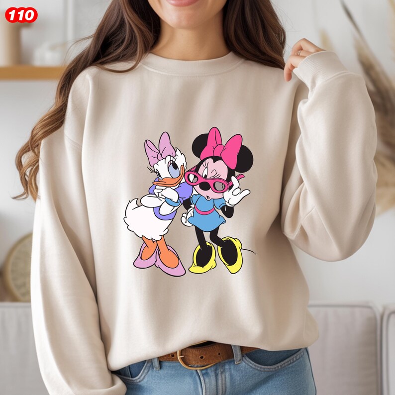 Pode incluir: Camisola bege com um gr&aacute;fico colorido da Minnie Mouse e da Margarida. Minnie Mouse usa um la&ccedil;o rosa e &oacute;culos de sol, enquanto Margarida usa um la&ccedil;o e &oacute;culos roxos. A camisola tem mangas compridas e gola redonda.