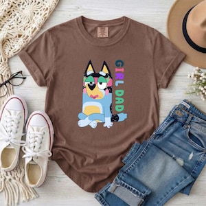 May include: A brown short-sleeved t-shirt featuring a graphic of a blue cartoon dog sitting with legs crossed. The dog has pink accents on its ears and face. The text GIRL DAD is printed vertically in colorful block letters next to the dog illustration.