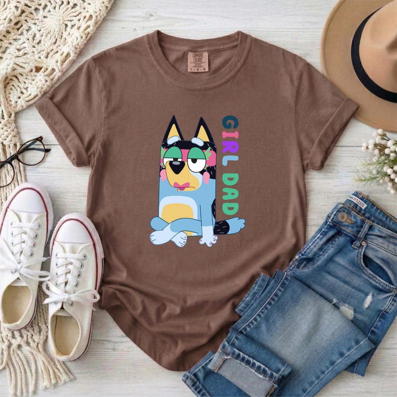 May include: A brown short-sleeved t-shirt featuring a graphic of a blue cartoon dog sitting with legs crossed. The dog has pink accents on its ears and face. The text GIRL DAD is printed vertically in colorful block letters next to the dog illustration.