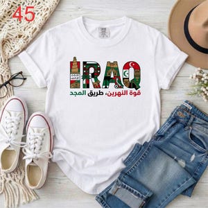 Puede incluir: Una camiseta blanca de manga corta con la palabra IRAQ en letras grandes, cada una rellena con ilustraciones coloridas de monumentos y s&iacute;mbolos culturales, con texto en &aacute;rabe impreso debajo en tinta verde y roja.