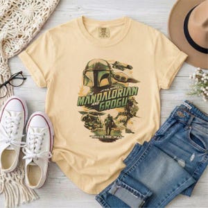 May include: A light yellow t-shirt featuring a graphic from "The Mandalorian" with the text "The Mandalorian and Grogu" and "This is the Way." The shirt is paired with blue jeans, white sneakers, and a tan hat.