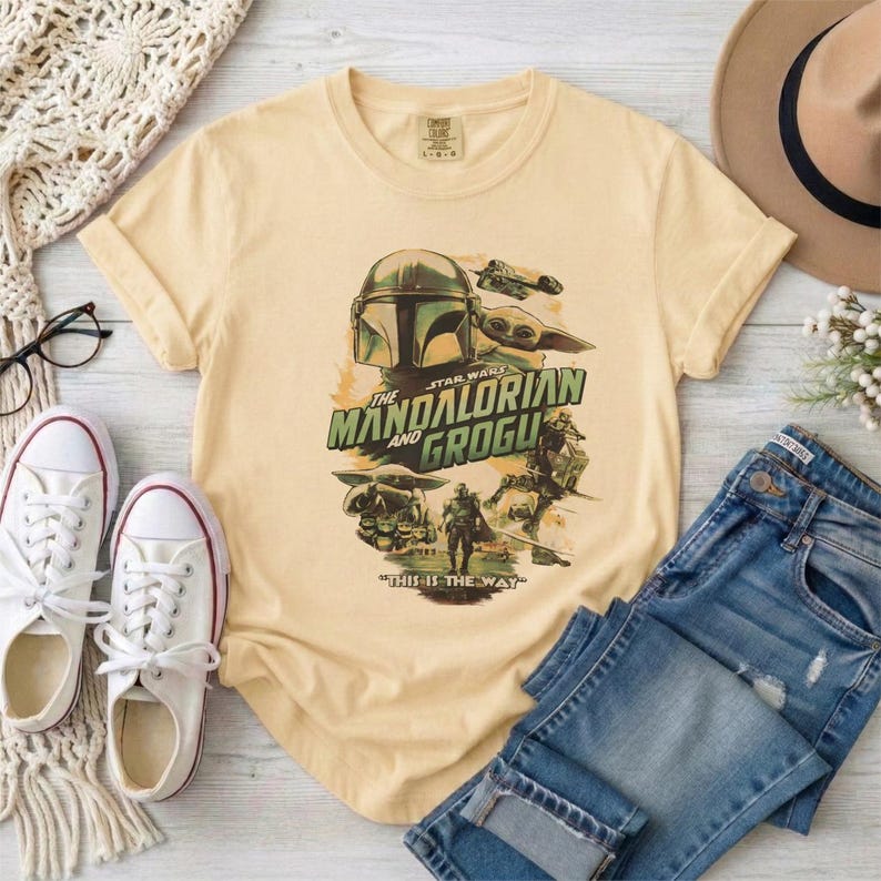 May include: A light yellow t-shirt featuring a graphic from "The Mandalorian" with the text "The Mandalorian and Grogu" and "This is the Way." The shirt is paired with blue jeans, white sneakers, and a tan hat.