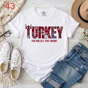 Puede incluir: Una camiseta blanca de manga corta con un dise&ntilde;o gr&aacute;fico que muestra la palabra TURKEY en letras rojas grandes rellenas de varios iconos y patrones culturales. Debajo de la palabra, el texto dice TEK MILLET, TEK TAKIM! en letras rojas.