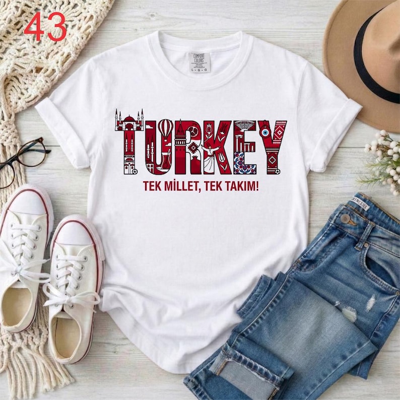 Puede incluir: Una camiseta blanca de manga corta con un dise&ntilde;o gr&aacute;fico que muestra la palabra TURKEY en letras rojas grandes rellenas de varios iconos y patrones culturales. Debajo de la palabra, el texto dice TEK MILLET, TEK TAKIM! en letras rojas.