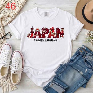 Puede incluir: Una camiseta blanca de manga corta con la palabra JAPAN en letras rojas grandes rellenas con ilustraciones de una pagoda, el monte Fuji, flores de cerezo, un abanico, un bons&aacute;i y un maneki-neko. Debajo aparece el texto: 日本の誇り、世界を驚かせ.