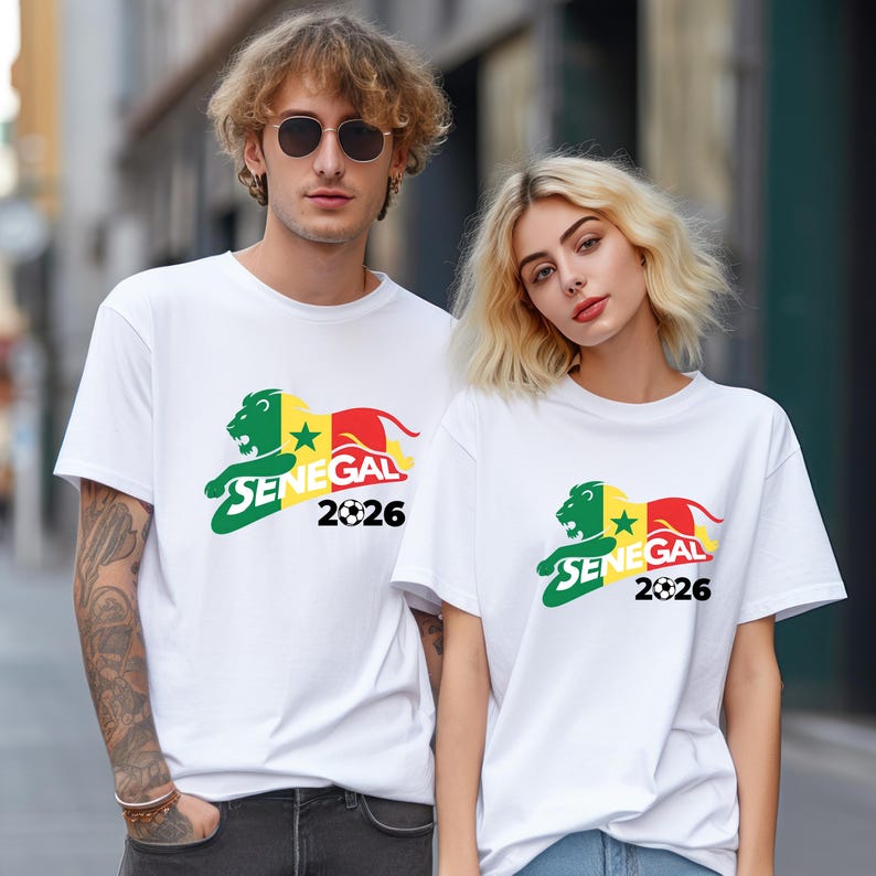 Pu&ograve; includere: Due persone indossano magliette bianche. Ogni maglietta mostra una grafica con un leone verde, una stella gialla e la parola "SENEGAL" in verde, con l'anno "2026" sotto. Il leone ha accenti rossi e gialli.
