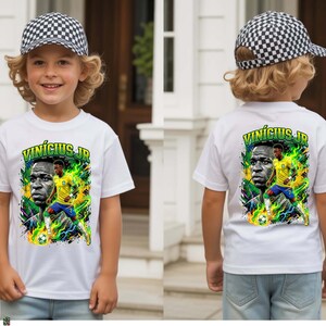 Puede incluir: Camiseta blanca con un gr&aacute;fico del futbolista Vin&iacute;cius J&uacute;nior con el uniforme de la selecci&oacute;n brasile&ntilde;a. El dise&ntilde;o incluye el retrato del jugador y su nombre en verde y amarillo. El ni&ntilde;o lleva una gorra a cuadros blancos y negros.