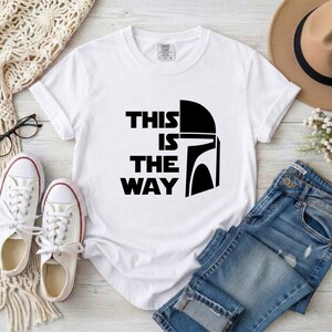 May include: White t-shirt with the text "THIS IS THE WAY" and a black helmet graphic. The shirt is paired with blue jeans, white sneakers, and a tan hat. A pair of glasses and a cream-colored knit scarf are also visible.