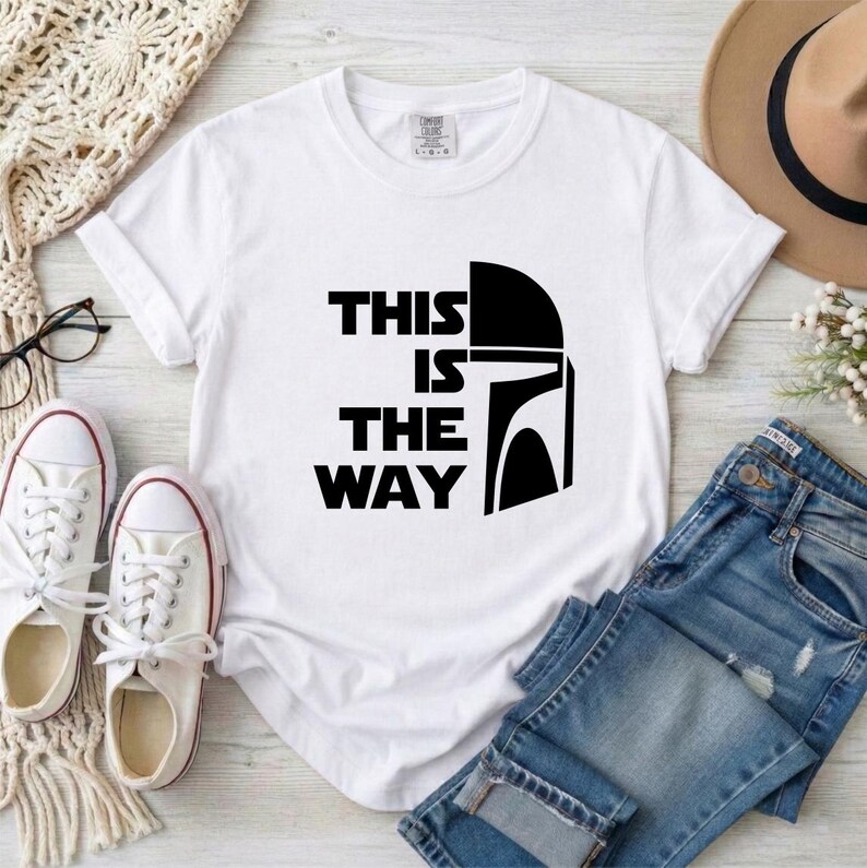May include: White t-shirt with the text "THIS IS THE WAY" and a black helmet graphic. The shirt is paired with blue jeans, white sneakers, and a tan hat. A pair of glasses and a cream-colored knit scarf are also visible.
