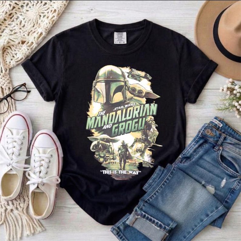 May include: Black t-shirt featuring a graphic from the Star Wars series, "The Mandalorian and Grogu." The design includes images of the Mandalorian, Grogu, and other characters, with the phrase "This is the Way" at the bottom. The shirt is paired with blue jeans and white sneakers.