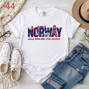 Puede incluir: Una camiseta blanca de manga corta con la palabra NORWAY en letras grandes azules y rojas, con ilustraciones detalladas en su interior. Debajo, el texto dice ALLE SOM &Eacute;N: FOR NORGE en letras azules y rojas.