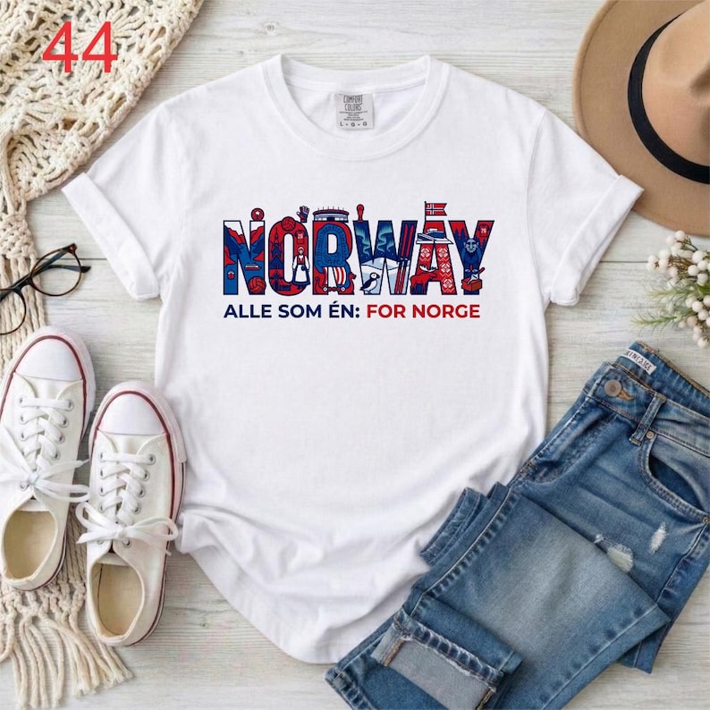 Puede incluir: Una camiseta blanca de manga corta con la palabra NORWAY en letras grandes azules y rojas, con ilustraciones detalladas en su interior. Debajo, el texto dice ALLE SOM &Eacute;N: FOR NORGE en letras azules y rojas.
