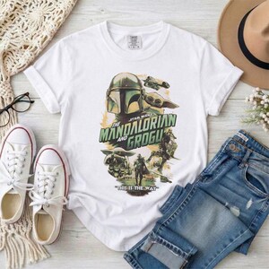 May include: White t-shirt featuring a graphic from "The Mandalorian and Grogu." The design includes the Mandalorian helmet, Grogu, and other characters. The text "THIS IS THE WAY" is also included. The shirt is paired with blue jeans and white sneakers.