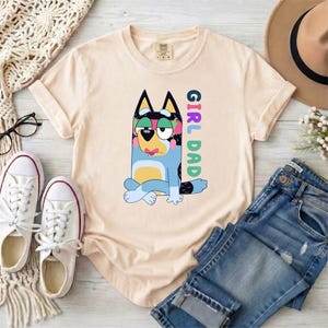 May include: A light beige short-sleeve t-shirt featuring a graphic of a blue and yellow cartoon dog sitting with legs crossed. The text "GIRL DAD" is printed vertically in colorful letters next to the dog. The shirt has a crew neckline.