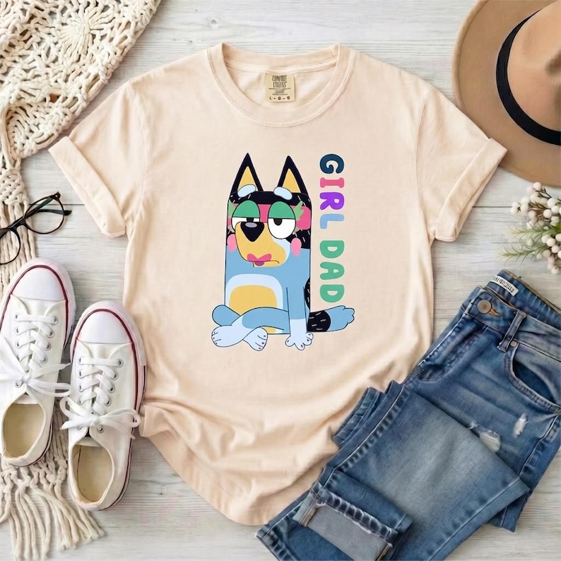 May include: A light beige short-sleeve t-shirt featuring a graphic of a blue and yellow cartoon dog sitting with legs crossed. The text "GIRL DAD" is printed vertically in colorful letters next to the dog. The shirt has a crew neckline.