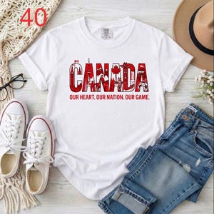 Puede incluir: Una camiseta blanca de manga corta con un dise&ntilde;o gr&aacute;fico rojo que deletrea CANADA, incorporando varios iconos canadienses dentro de las letras. Debajo del gr&aacute;fico, el texto OUR HEART. OUR NATION. OUR GAME. aparece en letras may&uacute;sculas rojas.