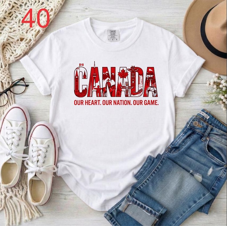 Puede incluir: Una camiseta blanca de manga corta con un dise&ntilde;o gr&aacute;fico rojo que deletrea CANADA, incorporando varios iconos canadienses dentro de las letras. Debajo del gr&aacute;fico, el texto OUR HEART. OUR NATION. OUR GAME. aparece en letras may&uacute;sculas rojas.