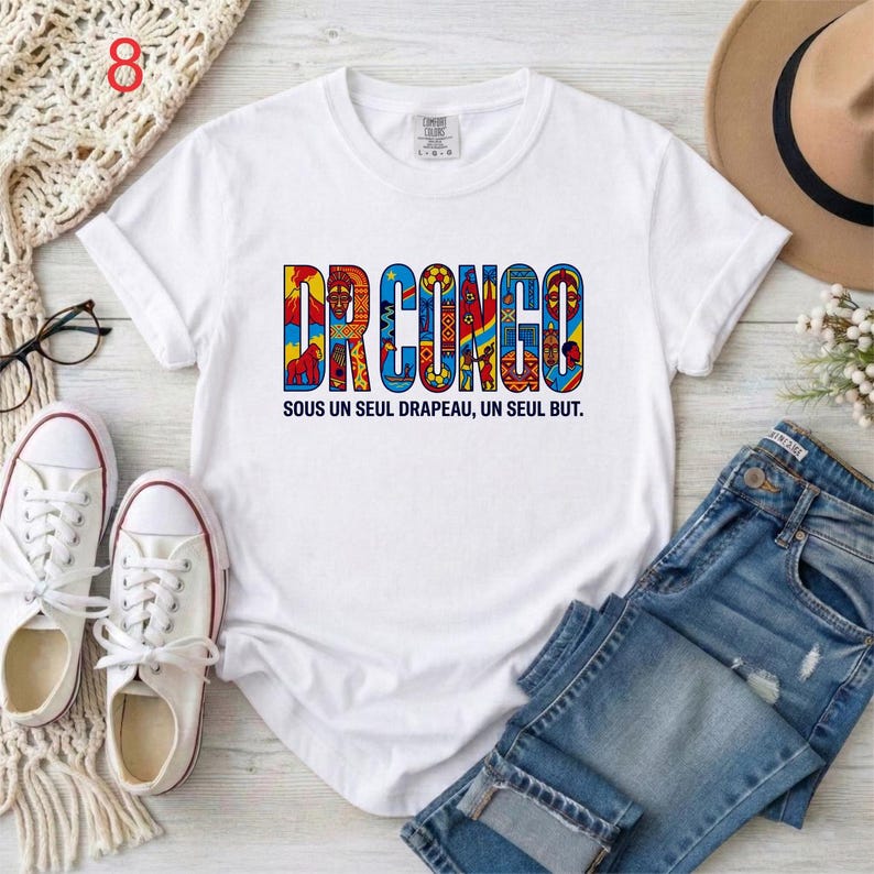 May include: White t-shirt with the text "DR CONGO" in colorful, patterned letters. Below the text is the phrase "SOUS UN SEUL DRAPEAU, UN SEUL BUT." The shirt is paired with blue jeans, white sneakers, and a beige hat.