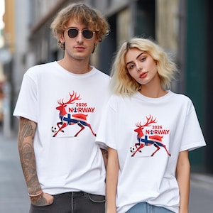 Pu&ograve; includere: Due persone indossano magliette bianche. Ogni maglietta presenta un disegno di renna rossa, la bandiera norvegese, un pallone da calcio e la scritta "2026 NORWAY". Le magliette sono a maniche corte.