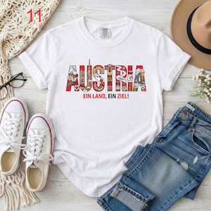 May include: White t-shirt with the word "AUSTRIA" in colorful letters, featuring Austrian landmarks and designs. Below the word is the phrase "EIN LAND, EIN ZIEL!" in red. The shirt is paired with blue jeans and white sneakers.