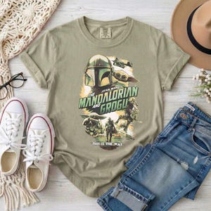 May include: Olive green t-shirt featuring a graphic from the Star Wars series, "The Mandalorian and Grogu." The design includes the Mandalorian helmet, Grogu, and the phrase "This is the Way."