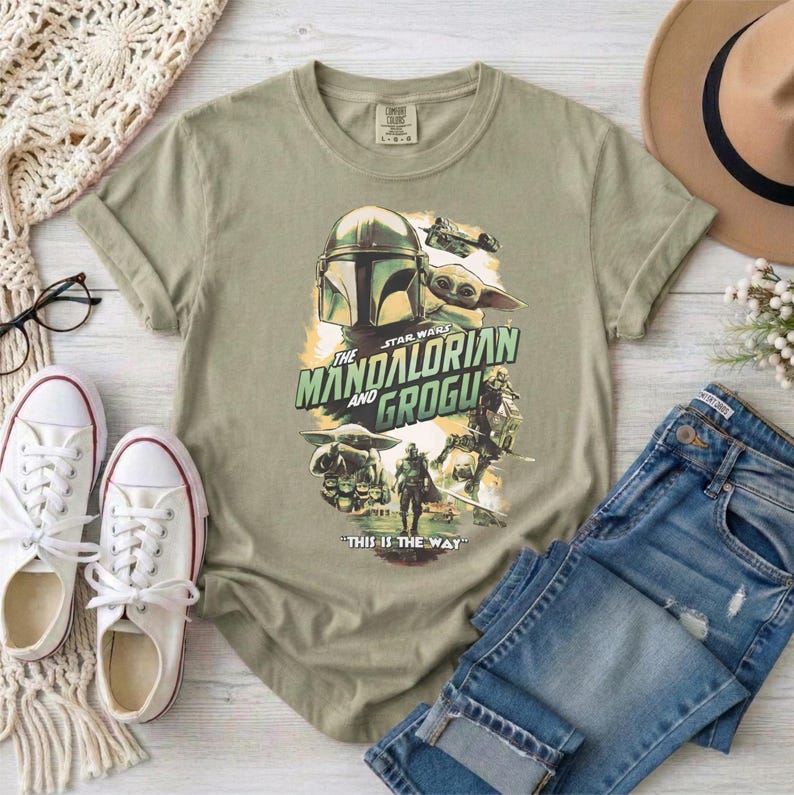 May include: Olive green t-shirt featuring a graphic from the Star Wars series, "The Mandalorian and Grogu." The design includes the Mandalorian helmet, Grogu, and the phrase "This is the Way."