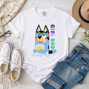 May include: A white short-sleeve t-shirt featuring a graphic of a blue cartoon dog with a yellow belly and pink accents. The dog is sitting with its legs crossed. To the right of the dog, the text "GIRL DAD" is printed vertically in colorful letters.