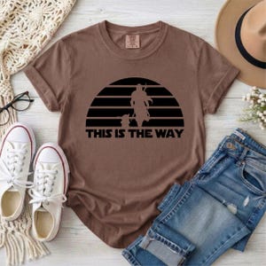 May include: Brown t-shirt with a black graphic of a silhouette of a figure and a child against a sunset design. The text "THIS IS THE WAY" is below the graphic. The shirt is paired with blue jeans and white sneakers.