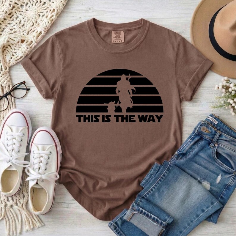 May include: Brown t-shirt with a black graphic of a silhouette of a figure and a child against a sunset design. The text "THIS IS THE WAY" is below the graphic. The shirt is paired with blue jeans and white sneakers.