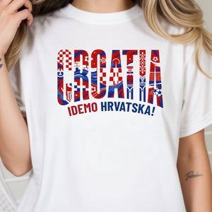 May include: White t-shirt with the word "CROATIA" in red, white, and blue, with various Croatian flag-inspired patterns filling each letter. Below the word is the phrase "IDEMO HRVATSKA!" in red. The shirt is a casual wear item.