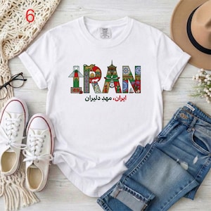May include: White t-shirt with the word "IRAN" in colorful, decorative letters. Below the word "IRAN" is text in a different language. The shirt is paired with blue jeans, white sneakers, and a brown hat.