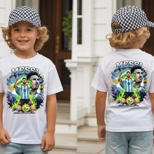 Puede incluir: Camiseta blanca con un gr&aacute;fico de un jugador de f&uacute;tbol y el nombre "MESSI" encima de la imagen. El jugador lleva una camiseta a rayas azules y blancas. El ni&ntilde;o tambi&eacute;n lleva una gorra de b&eacute;isbol a cuadros blancos y negros.