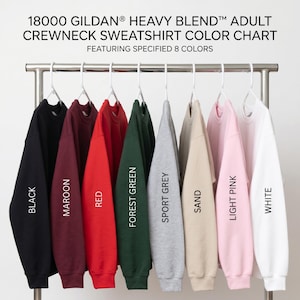May include: A Gildan 18000 Heavy Blend crewneck sweatshirt color chart featuring eight long-sleeve shirts hanging on white plastic hangers. The colors displayed from left to right are black, maroon, red, forest green, sport grey, sand, light pink, and white.