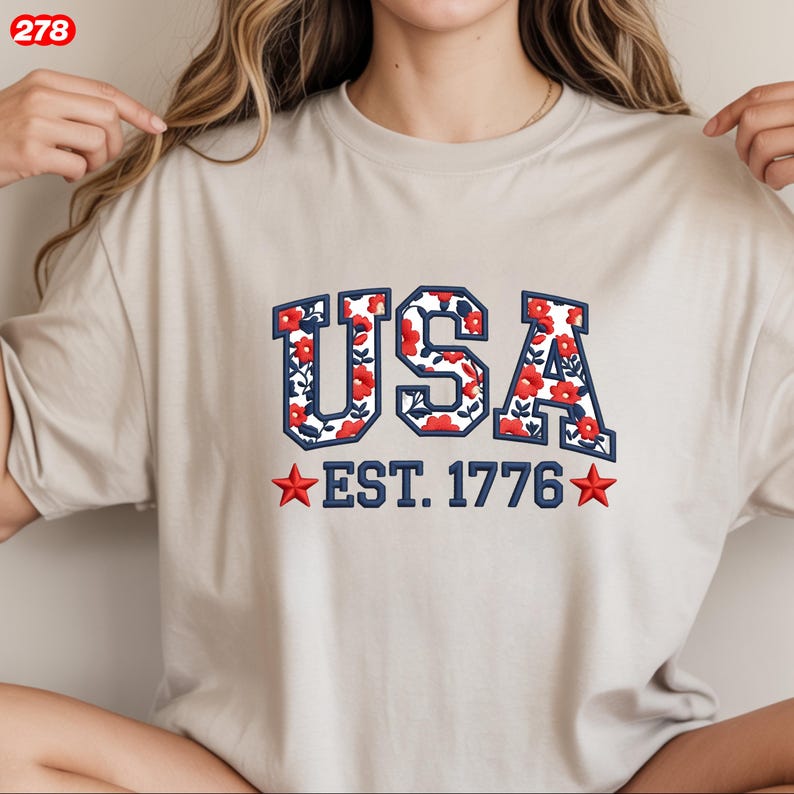 Pode incluir: Uma camiseta bege com a palavra "USA" em contorno azul marinho, preenchida com um padr&atilde;o floral de flores vermelhas e folhas verdes. Abaixo est&aacute; o texto "EST. 1776" com estrelas vermelhas em cada lado.